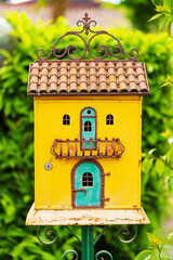 close-up of a colorful decorative birdhouse with a tiled roof, set in a garden with green foliage