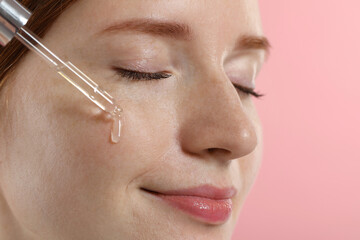 Obraz premium Beautiful woman with freckles applying cosmetic serum onto her face on pink background, closeup