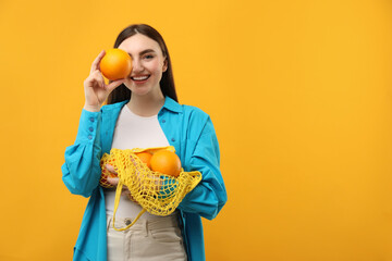 Woman holding string bag of fresh oranges and covering eye with fruit on orange background, space...