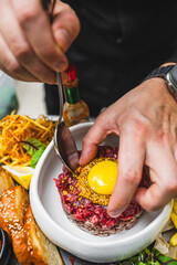 Close-up of hands preparing a gourmet dish, adding an egg yolk on top of seasoned raw minced meat, with garnishes around