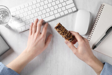 Woman holding tasty granola bar working with computer at light table, top view