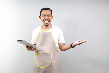 happy smiling asian man barista bartender barman employee wear brown apron white t-shirt work in coffee shop using tablet pc computer spread hand isolated on gray background. business startup - Powered by Adobe