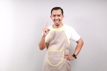 Happy Asian male housekeeper chef cook baker man wear apron showing a finger isolated on gray...