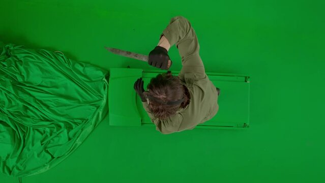 Maniac in hockey mask and jumpsuit running and waving a large sharp knife. An unrecognizable man in the terrifying guise of Jason Voorhees on a green screen chromakey. Halloween, horror. Top view.
