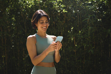 Happy Hispanic fit young woman using mobile phone checking fitness applications standing in green...