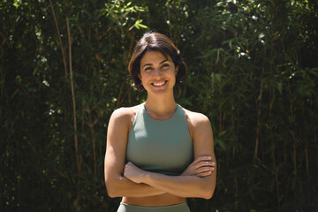 Happy fit sporty active slim confident young Hispanic woman wearing sportswear enjoying summer sunny day looking at camera standing arms crossed in green nature park. Portrait.