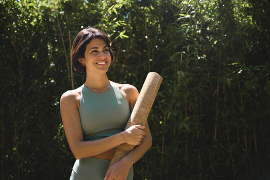 Happy fit confident young Hispanic woman advertising yoga retreat outdoor class. Sporty active lady wearing sportswear holding mat looking away standing in green nature park. Copy space for ads.