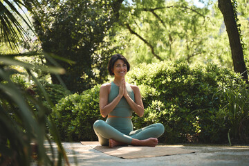 Happy mindful young Hispanic woman meditating doing yoga breathing exercises feeling peace of mind, mental balance sitting in lotus pose hands in namaste in green nature park outdoors.
