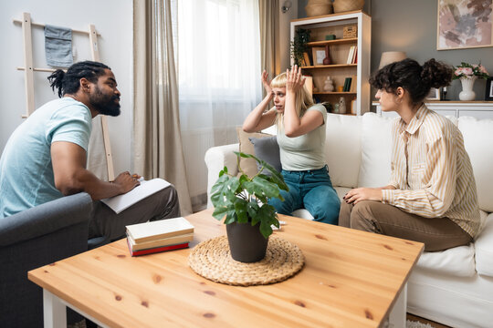 Stressed African American male psychologist and angry LGBT lesbian couple fighting during session at doctors office. Homosexual spouses having conflict on consultation with psychotherapist.