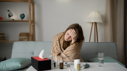young sick woman sits on the sofa at home, bends over a table with pills and holds her head with...