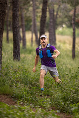 A man is running through a forest wearing a purple shirt and gray shorts. He is wearing a hat and a backpack
