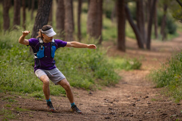 A man in a purple shirt and gray shorts is playing frisbee in a forest. He is wearing a visor and a...