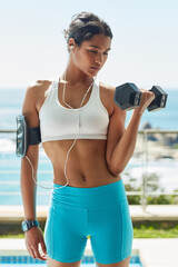 Indian girl, earphones and dumbbell for workout in outdoor, bicep and training for muscle growth. Female person, confident and music for weight lifting, strong body and sound for power or health