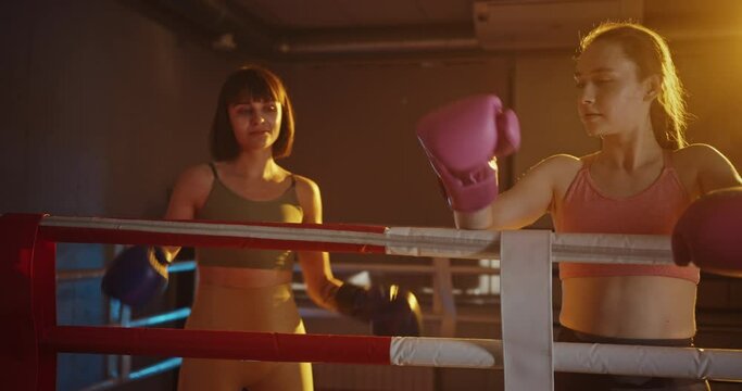 Two young female boxers rest after an intense training session in a boxing ring, showcasing resilience and determination under warm, atmospheric lighting