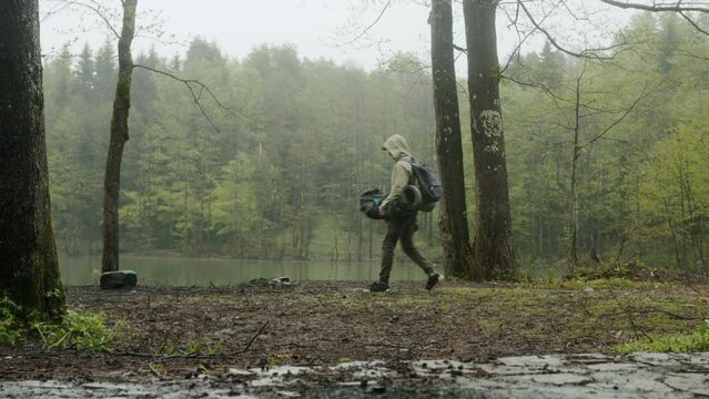 Hiker man assembles campsite tent in the rainy forest near the lake. Traveler installing tent in woods. Outdoor weekend activity. Wide shot