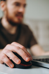 A male hand holds a computer mouse near a laptop on the background of a man working at a computer.