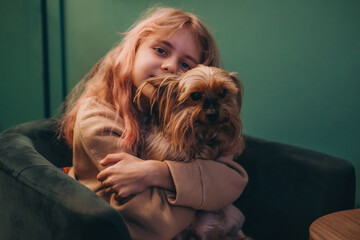 A beautiful little girl hugs a Yorkshire terrier while sitting on a chair.