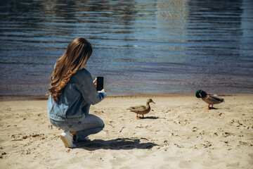 A young woman takes pictures of ducks on the riverbank on her phone.