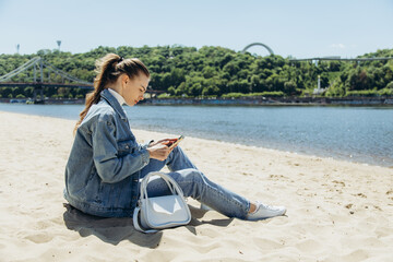 A young woman sits on the sand at the beach and looks at the phone.