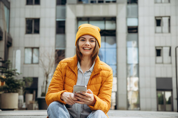 Happy smiling woman with braces in yellow jacket sitting on the street and holds the phone in her hands.