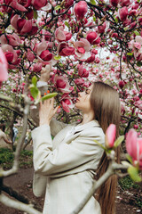 Fototapeta premium A young woman in a jacket enjoys blooming magnolia trees under a large tree.