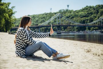 A young woman sits on the sand at the beach and smiling at the phone.