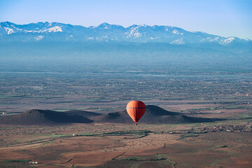 A red, hot air balloon, floating over the desert near Marrakech, Morocco on a clear spring day, with the snow capped Atlas mountains in the background © parkerspics