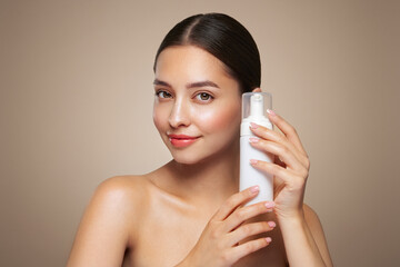 A young woman shows off a bottle with a foam dispenser for washing. Daily skin care cosmetics advertisement. Studio shoot Beige background