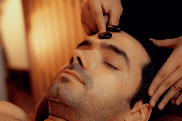 Caucasian man enjoying relaxing anti-stress head massage with hot stone and pampering facial beauty...