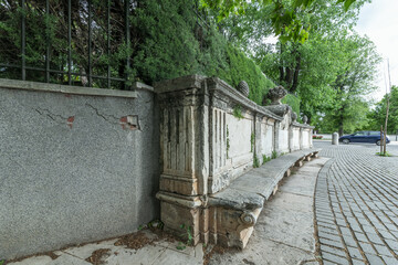 An ornate antique stone bench in a park next to a hedge with stone cobblestone floors