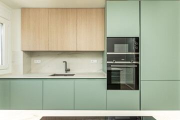 Front image of a modern design open kitchen with pastel green furniture, white circular decorative hood, columns of integrated appliances and marble backsplash