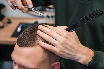 Hairstylist carefully cuts the brunette hair of the client with scissors at the barbershop. 