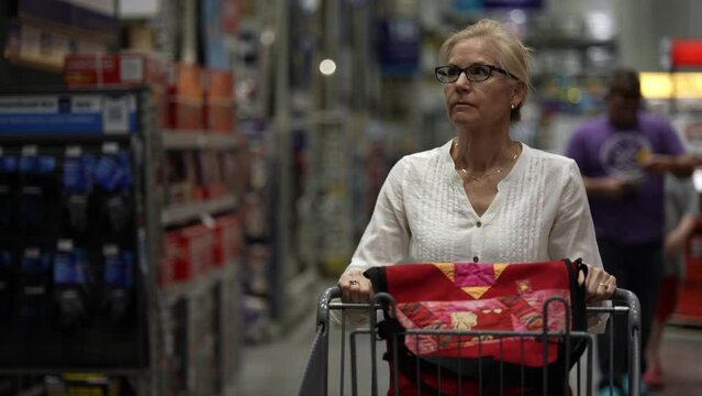 Closeup front view of mature woman pushing shopping cart in home security section of a big box hardware store shopping for home renovation construction project.