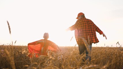 Father farmer and little daughter superhero in red cloak running at sunset wheat field. Happy...