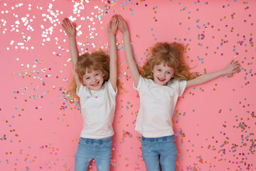 Twin girls celebrate birthday. Smiling, positive curly blonde sisters 6 years old, dressed in white T-shirts and jeans, lie on a pink background with confetti and look at the camera and holding hands.