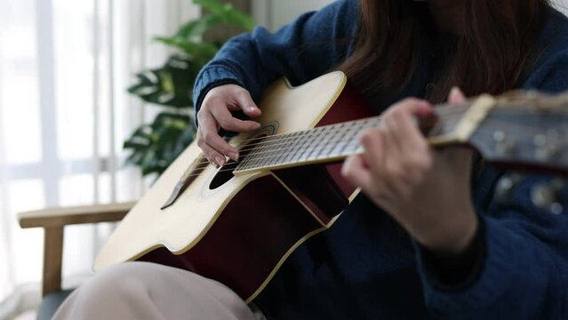 Asian girl playing acoustic guitar in living room. Happy songwriter playing chords on a guitar-inspired string instrument playing instrument chords at home.