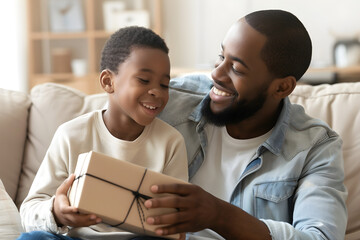 Little black son surprising his African American father with a father’s day gift while sitting together on the sofa at home.