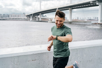 A man checks his watch while jogging beside a waterfront, with a modern bridge in the background.
