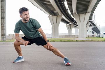 A man stretches his leg under a large bridge, preparing for a workout session on a clear day.