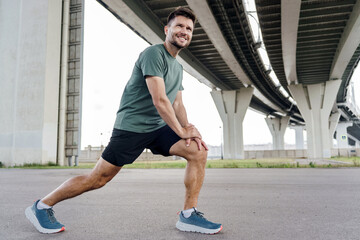 A man stretches his leg under a bridge, preparing for an outdoor workout session with a smile.