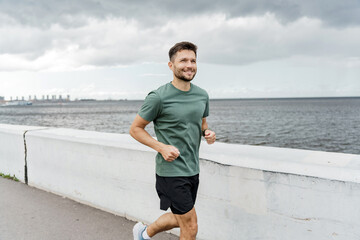 A man jogs along a waterfront pathway, with a scenic water view and cloudy sky in the background.