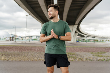 A man stands under a bridge, smiling and clasping his hands, preparing for his workout session