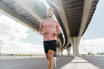 A man jogs under a large bridge, smiling as sunlight filters through, highlighting his athletic form.