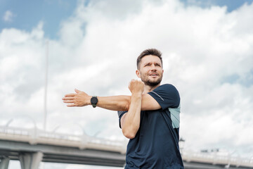 A man performs an arm stretch while standing outdoors, with a bridge and cloudy sky in the background