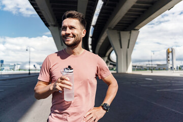 A man smiles while holding a water bottle under a bridge, enjoying a break during his workout.