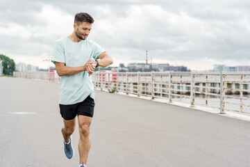 A man checks his watch while running on a waterfront path, with a cloudy sky in the background.