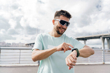 Man in sunglasses checking his smartwatch, smiling, wearing a light blue shirt, by a waterfront.