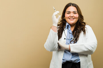 Good looking plus-size female doctor holding a syringe, posing confidently in a studio with copy...
