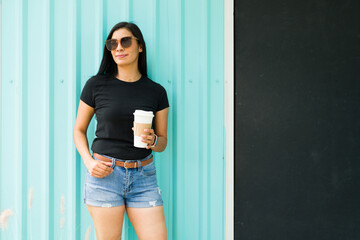 Self-assured latina woman in a black t-shirt and sunglasses holding a coffee cup with a bright...