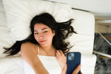 Young woman lying on a bed in a sunlit room, taking a sexy selfie with her smartphone